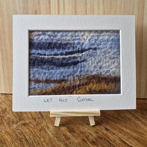 May include: A framed wet felt coastal landscape art piece. The artwork features a textured design with shades of blue, brown, and white, evoking a seaside scene. The white frame is displayed on a small wooden easel.