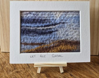 May include: A framed wet felt coastal landscape art piece. The artwork features a textured design with shades of blue, brown, and white, evoking a seaside scene. The white frame is displayed on a small wooden easel.