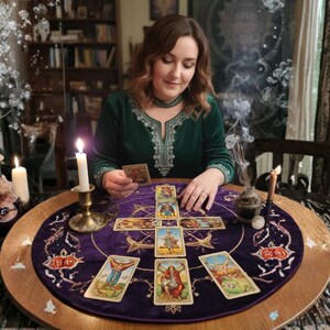 May include: A woman in a green velvet dress is reading tarot cards on a round table. The table is covered with a purple cloth with a tarot card layout. Lit candles and incense add to the mystical atmosphere. The scene is set in a room with a bookshelf.