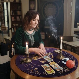 May include: A woman in a green velvet dress sits at a round table with tarot cards, lit candles, and a small pumpkin. The table is covered with a purple mat with star and moon designs. Incense smoke rises in the air.
