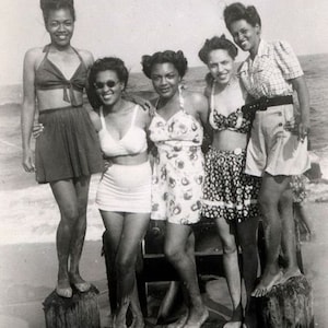 Puede incluir: Fotografía vintage en blanco y negro de cinco mujeres en trajes de baño posando sobre postes de madera en la playa. Las mujeres visten trajes de baño y pantalones cortos, con varios patrones y estilos. La imagen captura un momento de ocio y camaradería.