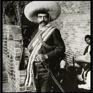 Puede incluir: Un hombre con un sombrero de ala ancha y uniforme militar sostiene un rifle y una espada. Está de pie frente a una pared de ladrillo. Otros dos hombres están de pie detrás de él.