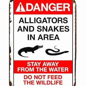 Peut inclure: Panneau métallique rectangulaire avec fond blanc et bordures rouges. Il indique "DANGER ALLIGATORS AND SNAKES IN AREA" avec des illustrations d'un alligator et d'un serpent. Le texte supplémentaire avertit de rester à l'écart de l'eau et de ne pas nourrir la faune.