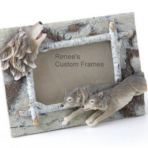 May include: A rectangular picture frame with a rustic design. The frame features a wolf howling at the top left corner and two running wolves at the bottom right. The frame is made of a light-colored material with a birch bark texture. The text "Renee's Custom Frames" is visible.
