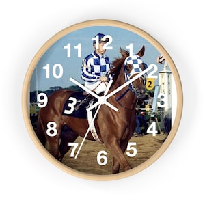 May include: Round wall clock with a light wood frame. The clock face features a vintage image of a jockey on a brown horse during a race. The clock hands are white, and the numbers are large and easy to read.