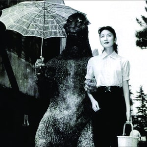 May include: A black and white photo of a woman standing next to a large, dinosaur-like creature. The creature is holding a white umbrella. The woman is wearing a white shirt and a dark skirt. She is holding a bucket in her right hand.