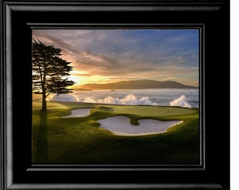 Puede incluir: Una fotografía panorámica de un campo de golf con un fairway verde, un bunker de arena y un árbol solitario en primer plano. El fondo presenta una masa de agua con olas rompiendo y una cadena montañosa en la distancia. El cielo es de un naranja y rosa vibrantes, lo que sugiere una puesta de sol.