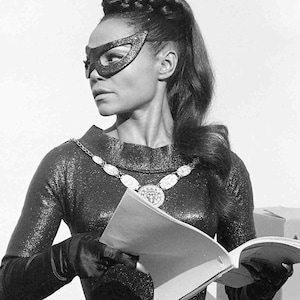 May include: A woman wearing a black catsuit with a sparkly texture and a black cat mask. She has a long, dark braid and is holding a script in her hands. She is standing in front of a white background.