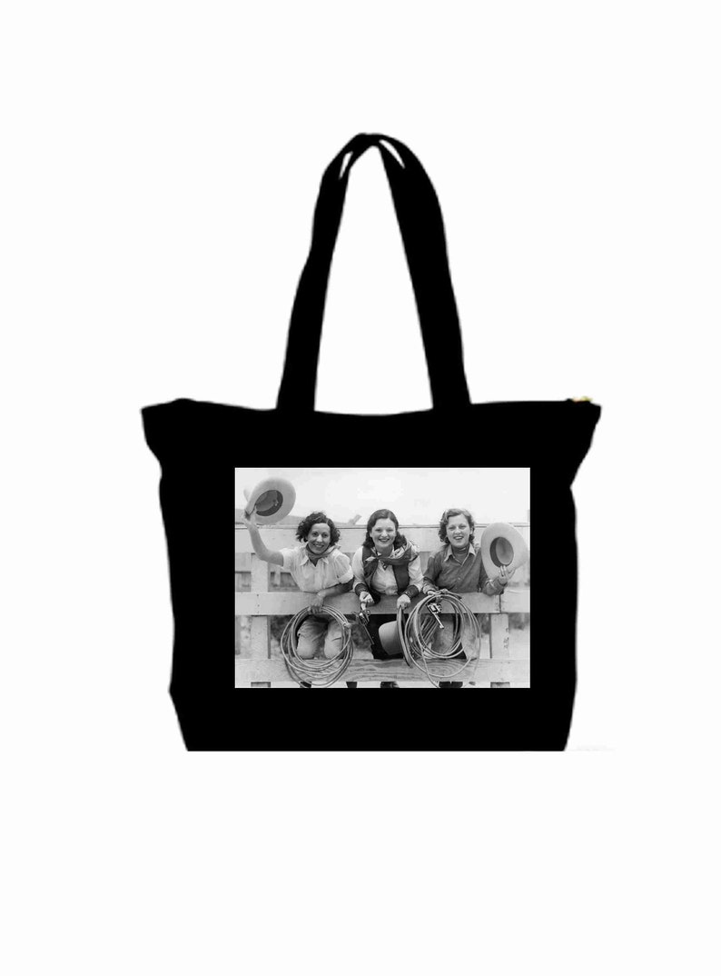 May include: Black and white photo of three women wearing cowboy hats and holding ropes. The women are sitting on a fence and smiling. The photo is printed on a black tote bag.