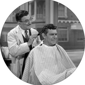 Puede incluir: Una fotografía en blanco y negro de un hombre que se corta el pelo con un barbero. El barbero lleva una bata blanca y sostiene un peine y unas tijeras. El hombre lleva una capa de barbero a rayas.