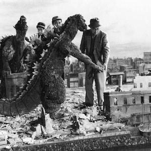 May include: A black and white photo of a large, scaly monster, possibly a dinosaur, being filmed on a movie set.  The monster is surrounded by people, some of whom are wearing hats and coats.  The set appears to be a destroyed city.