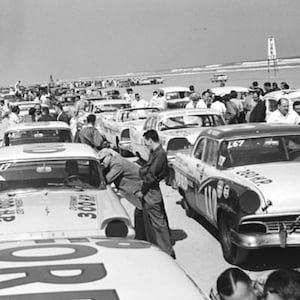 Puede incluir: Una fotografía en blanco y negro de una carrera de coches antiguos. La foto muestra una línea de coches clásicos, algunos con números de carrera, y una multitud de personas viendo la carrera. Los coches están alineados en una playa con el océano de fondo.
