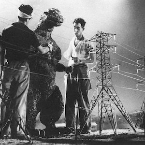 May include: A black and white photo of a man in a suit standing next to a large, scaly monster. The monster is standing in front of a row of power lines.