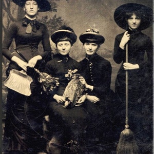 May include: Four women dressed in black clothing and witch hats pose for a vintage photograph. Two women hold brooms, one woman holds an owl, and one woman holds a fan.