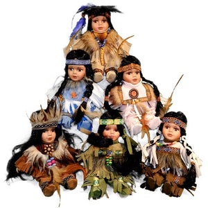 Puede incluir: Seis muñecas de porcelana con temática nativa americana. Cada muñeca viste atuendos tradicionales con intrincados abalorios, plumas y flecos. Las muñecas tienen cabello oscuro y están dispuestas en forma de pirámide.