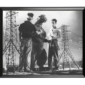 May include: Black and white framed print of a scene from a classic monster movie. Two men stand near a Godzilla-like creature, with miniature power lines and towers in the background.