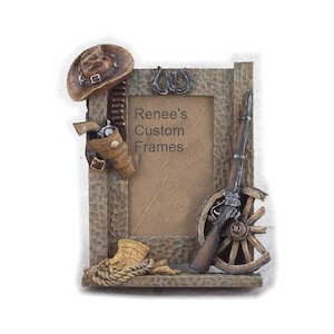 May include: A rustic wooden picture frame with a cowboy theme. It features a cowboy hat, a gun holster, a horseshoe, and a wagon wheel. The frame is decorated with a textured wood finish. The text "Renee's Custom Frames" is printed on the frame.