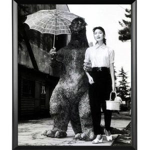 May include: Black and white framed photograph featuring a woman and a Godzilla figure. The Godzilla figure holds an umbrella. The woman is wearing a white blouse, black pants, and white shoes. She is holding a white basket.
