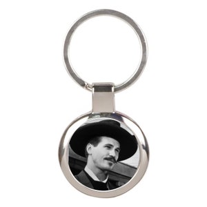 May include: A silver keychain with a round, black and white photo of a man wearing a hat. The man has a mustache and is looking to the right.