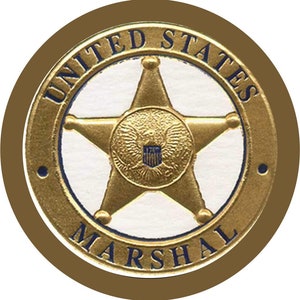 Puede incluir: Una insignia dorada de United States Marshal con una estrella de cinco puntas en el centro. Las palabras "UNITED STATES MARSHAL" están inscritas alrededor de la estrella en un patrón circular. La insignia tiene un borde marrón.