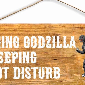 Puede incluir: Un letrero de madera con el texto "WARNING GODZILLA IS SLEEPING DO NOT DISTURB" en pintura blanca. Un monstruo Godzilla negro y gris se representa en el lado derecho del letrero.