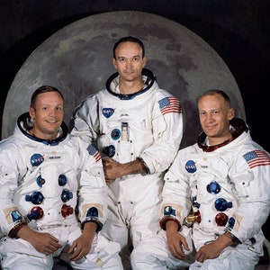 May include: Three astronauts in white spacesuits with NASA patches and American flags on their chests, sitting in front of a large, gray, moon-like sphere.