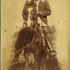 May include: A black and white photograph of a Native American man wearing a feathered headdress and a jacket, riding a horse. The man is looking directly at the camera. The photograph is faded and has a vintage look.