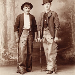 May include: Two men in vintage clothing stand in front of a backdrop of trees. The man on the left is wearing a dark jacket, a white shirt, and brown leather pants with fringe. The man on the right is wearing a tweed jacket, a dark shirt, and light brown pants with fringe. Both men are wearing hats.