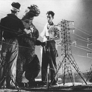 May include: A black and white photo of a man in a black shirt and a man in a white shirt standing next to a large, gray, dinosaur-like creature. The creature is standing in front of a set of power lines.