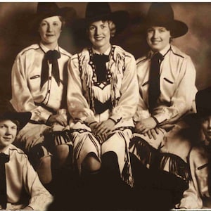 May include: Five women wearing cowboy hats and western-style shirts. The women are all smiling and looking at the camera. They are wearing different coloured shirts and hats.