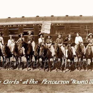 May include: A group of women in Western attire riding horses in front of a train. The women are wearing hats and long skirts. The train is a Southern Pacific train. The sign on the train says "Queen of the Round-Up". The women are smiling and looking at the camera. The image is in black and white.