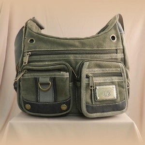 Può includere: Una borsa a tracolla in tela verde con più tasche con cerniera e una tasca con patta. La borsa ha una tracolla grigia e un'emblema metallico sulla tasca anteriore. La borsa è progettata con un'estetica casual e utilitaria.