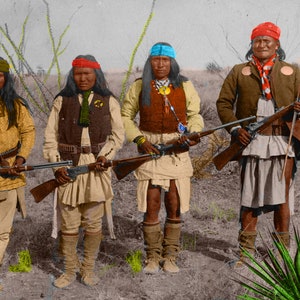 Könnte beinhalten: Vier indianische Männer stehen in einer Wüstenlandschaft, jeder mit einem Gewehr in der Hand. Sie tragen traditionelle Kleidung und Stirnbänder. Die Männer schauen direkt in die Kamera.