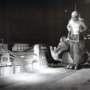 May include: A black and white photo of a man in a white hat operating a large, furry, monster puppet. The puppet is standing in front of a miniature city set.