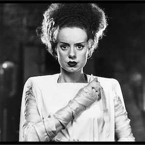 Pode incluir: Uma fotografia a preto e branco de uma mulher com um penteado que lembra o monstro de Frankenstein. Ela está vestida com um vestido branco e tem uma expressão séria no rosto.