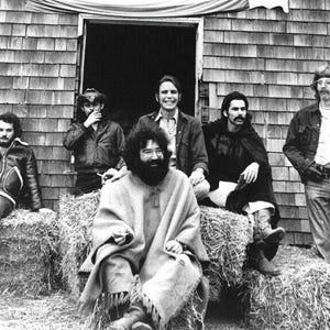 May include: Black and white photo of a group of six men standing in front of a wooden building. The men are wearing casual clothing and are smiling. They are standing in front of a pile of hay.