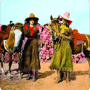Puede incluir: Fotografía vintage coloreada de dos mujeres con atuendo del oeste, cada una de pie junto a un caballo. Llevan sombreros, faldas con flecos y chaquetas. El fondo presenta una estructura de piedra rojiza y un cielo azul claro.