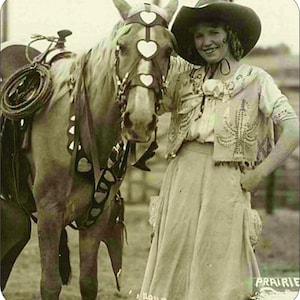 Puede incluir: Una joven con un vestido blanco y un sombrero de vaquero está de pie junto a un caballo marrón. El caballo lleva una brida decorada con corazones. La mujer sonríe y sostiene las riendas del caballo.