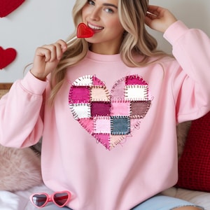 Puede incluir: Una sudadera rosa claro con un diseño de corazón de retazos en tonos de rosa, blanco y burdeos. El corazón está hecho de cuadrados de tela cosidos. La sudadera tiene cuello redondo y mangas largas.