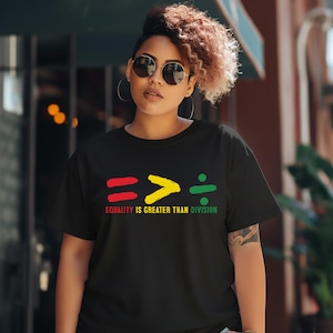 May include: Black t-shirt with the slogan "Equality is Greater Than Division" in red, yellow, and green. The design includes a mathematical symbol. The shirt is worn by a person with curly hair and sunglasses.