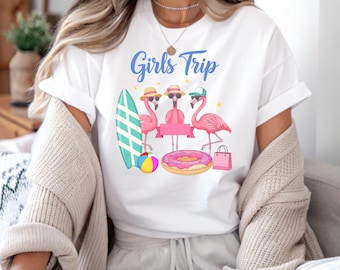 May include: White t-shirt with the words "Girls Trip" in blue. The design shows three pink flamingos wearing sunglasses and hats, alongside a surfboard, beach ball, a donut ring, and a shopping bag.