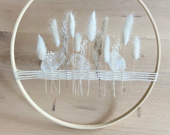 May include: A circular wall hanging featuring dried white flowers and grasses arranged within a wooden embroidery hoop. The design includes a woven section of twine and a hanging loop at the top. The natural elements create a minimalist, bohemian aesthetic.