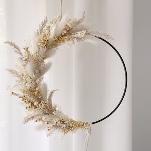 May include: A circular wall hanging featuring dried flowers and grasses in shades of cream and beige. The floral arrangement is attached to a black metal hoop, creating a modern, minimalist design. The wreath is suspended by a white cord.