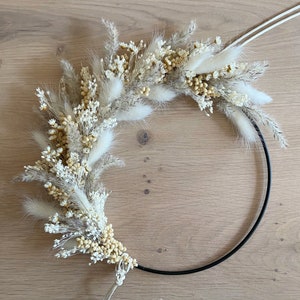 May include: A decorative wreath featuring dried flowers and grasses in shades of cream, beige, and tan. The floral arrangement is attached to a black metal ring, creating a modern, minimalist design. The wreath is perfect for adding a touch of natural beauty to any space.