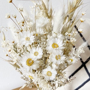May include: A bouquet of dried white flowers with yellow centers, including daisies and pampas grass, arranged in a clear glass vase with a brown twine bow.