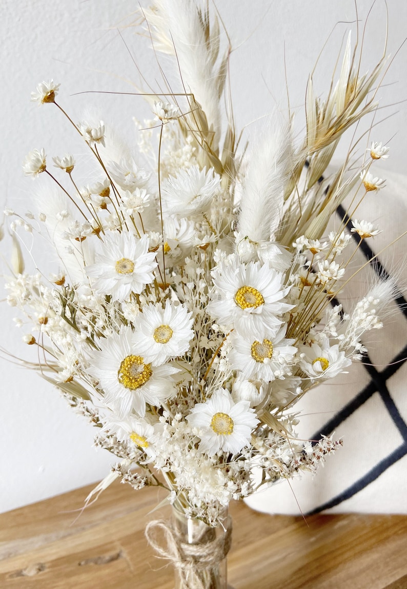 May include: A bouquet of dried white flowers with yellow centers, including daisies and pampas grass, arranged in a clear glass vase with a brown twine bow.
