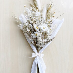 May include: A bouquet of dried white flowers, including daisies and baby's breath, wrapped in white tissue paper and tied with a white ribbon.