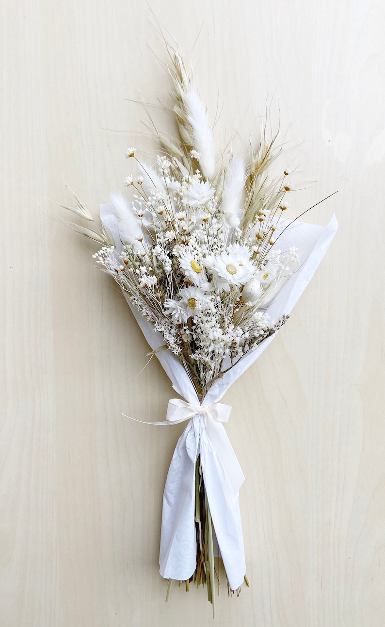 May include: A bouquet of dried white flowers, including daisies and baby's breath, wrapped in white tissue paper and tied with a white ribbon.