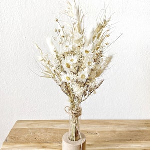 May include: A bouquet of dried white flowers and grasses arranged in a clear glass test tube vase, sitting in a wooden base.