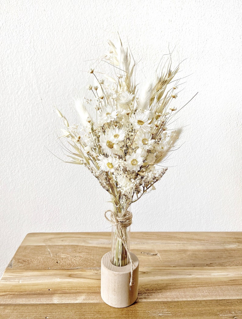 May include: A bouquet of dried white flowers and grasses arranged in a clear glass test tube vase, sitting in a wooden base.